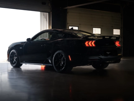 Rear three-quarter view of a black Ford Mustang in a dimly lit garage, highlighting the taillights and rear design.