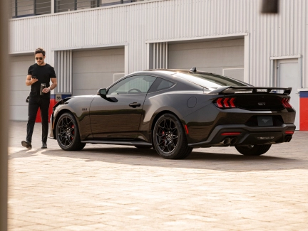 Black Ford Mustang side view parked next to a garage, person standing next to the vehicle.