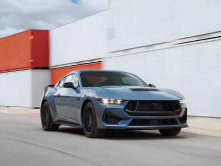 Blue Mustang driving in front of a modern building, showcasing its powerful stance.