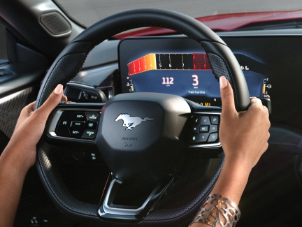 A hand on the Mustang's steering wheel, interacting with the cruise control buttons, demonstrating adaptive cruise control features.