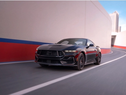 A dark gray Mustang shown from the front, driving along an urban road with high walls on the left.